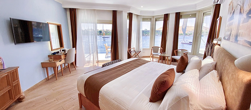 A bright corner room with floor-to-ceiling windows and river views, furnished with a soft bed and simple wooden accents.