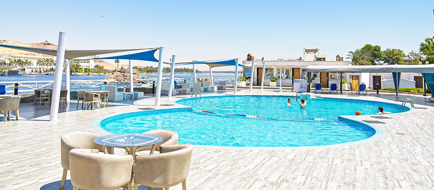 A bright pool terrace beside the Nile where families swim and relax under shade sails on a clear summer day.