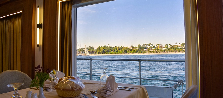 A dining table set beside a large picture window framing serene Nile views with passing feluccas.