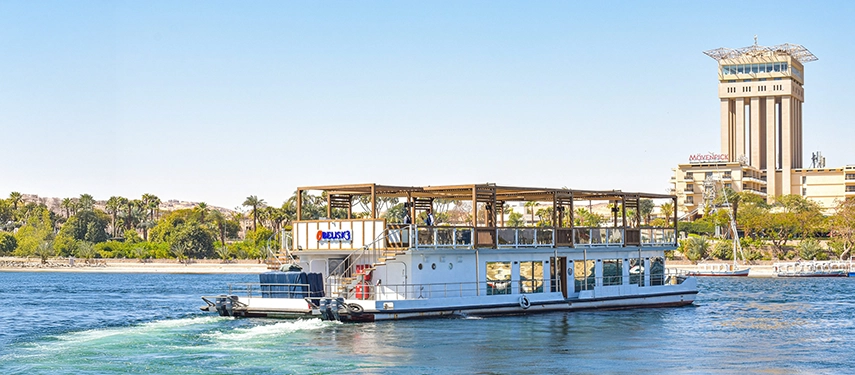 A Nile cruiser departing the riverbank with Aswan’s skyline and waterfront palms stretching out behind it.
