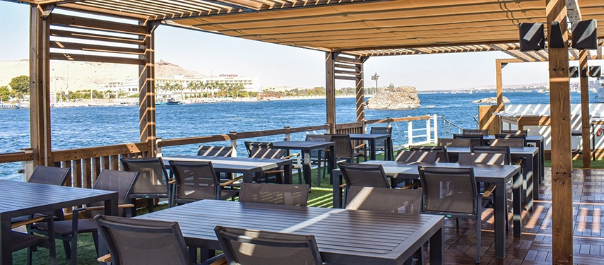 An al fresco riverside dining terrace shaded by wooden slats, overlooking the wide, calm waters of the Nile.