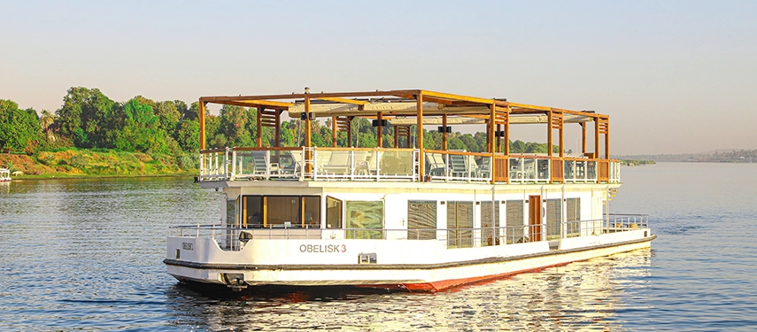 A bright white Nile cruiser moving slowly along the river, its open upper deck framed by timber beams.