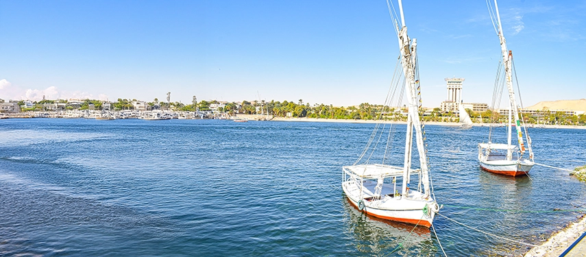 Two feluccas resting on the calm Nile with Aswan’s waterfront and desert hills stretching beneath a clear blue sky.