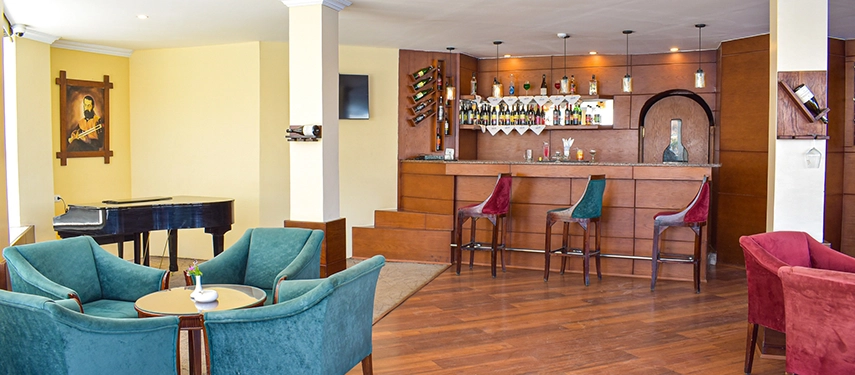 A warm, wood-panelled bar lounge with velvet chairs, a grand piano and a welcoming counter set for evening drinks.
