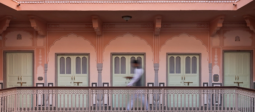A corridor of pastel pink walls and mint green doors, with a blurred figure walking past ornate arches.