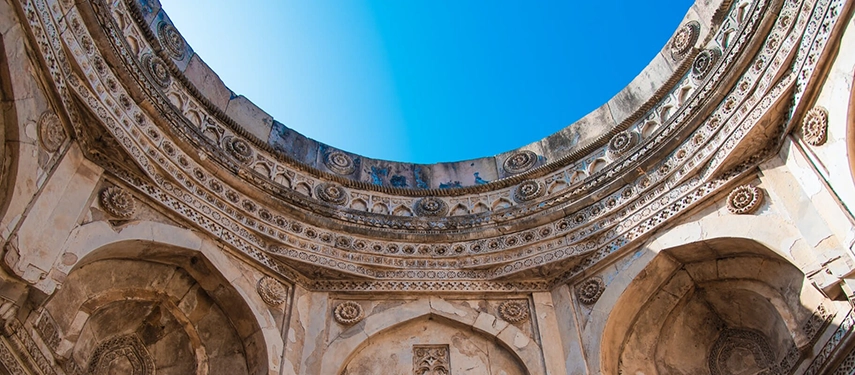 A dramatic upward view of an ancient dome interior with intricate carvings and a brilliant blue sky overhead.