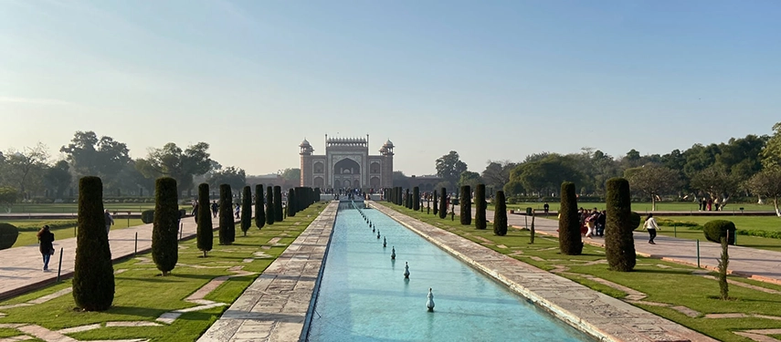 A long water channel flanked by manicured lawns and cypress trees, leading to an ornate Mughal-style gateway.