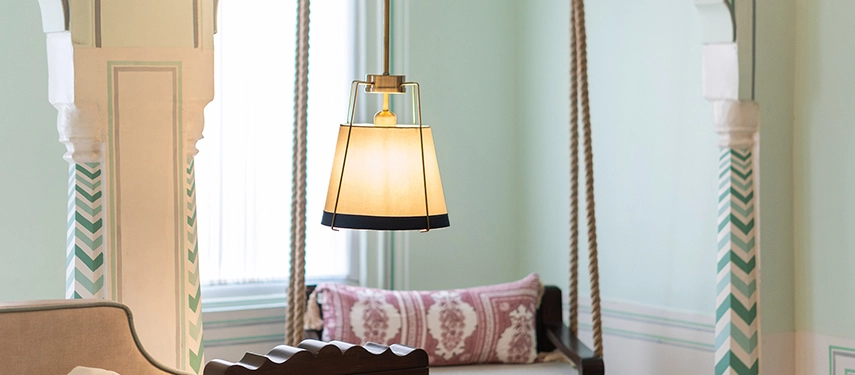 A close-up of a hanging pendant light with pastel green walls, carved arches, and a traditional wooden daybed in the background.