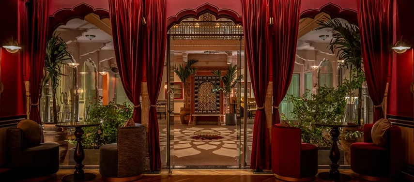 A romantic interior courtyard with red velvet curtains, potted palms, and ambient lighting at night.