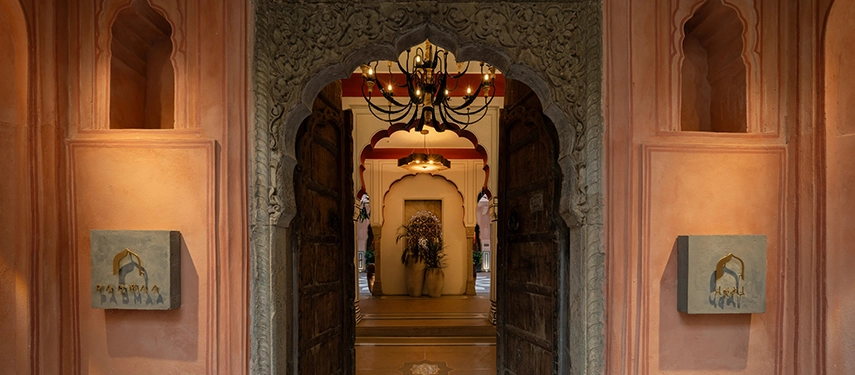 An intricately carved stone doorway with antique wooden doors leading into the warmly lit interior of Padmaa Haveli.