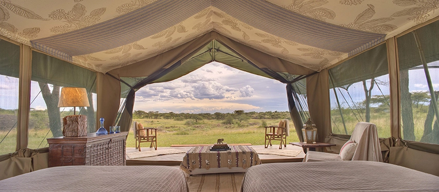 A spacious safari tent interior with twin beds, patterned canvas ceiling and open sides revealing a sweeping view across the Mara plains under a dramatic cloudy sky.