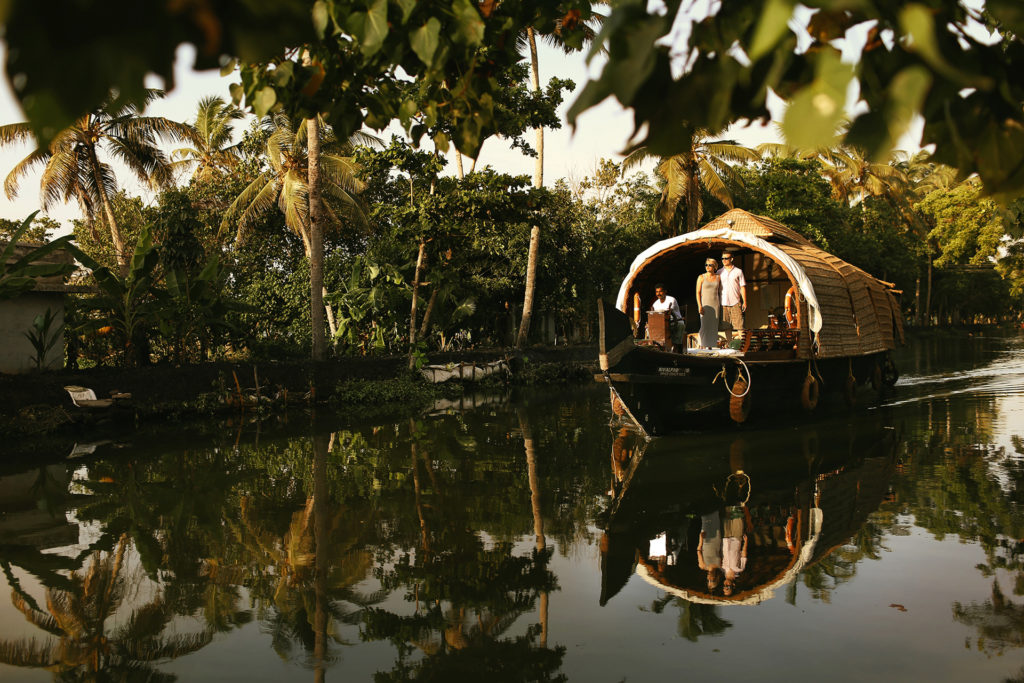 A traditional Kerala houseboat with a woven cane roof glides along a tranquil backwater canal, guests and crew visible on deck, the boat perfectly reflected in the still water below, Kerala, India.