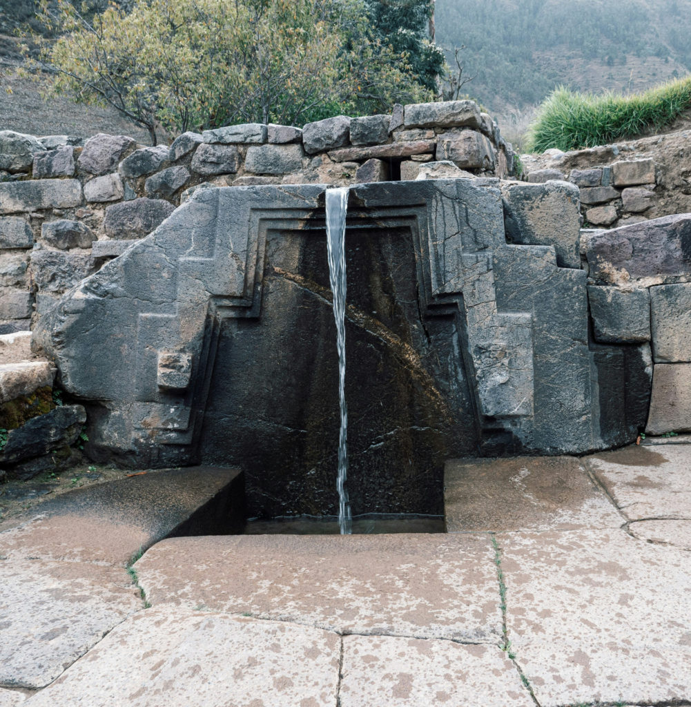 A carved ceremonial Inca stone fountain at Ollantaytambo, water flowing in a thin stream down an intricately stepped granite face into a stone basin below, Sacred Valley, Peru.