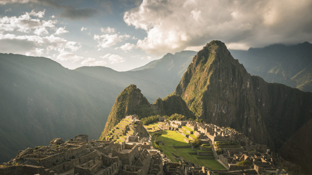 The iconic panoramic view of Machu Picchu at dusk, golden light illuminating the terraced citadel and green central plaza, Huayna Picchu mountain rising steeply behind under dramatic clouds, Peru.
