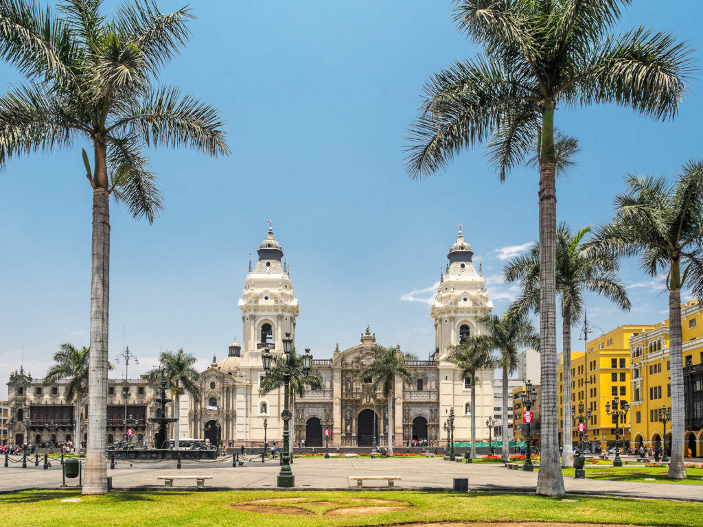 The white baroque façade of Lima Cathedral flanked by twin bell towers, seen across the palm-lined Plaza Mayor under a bright blue sky, Lima, Peru.