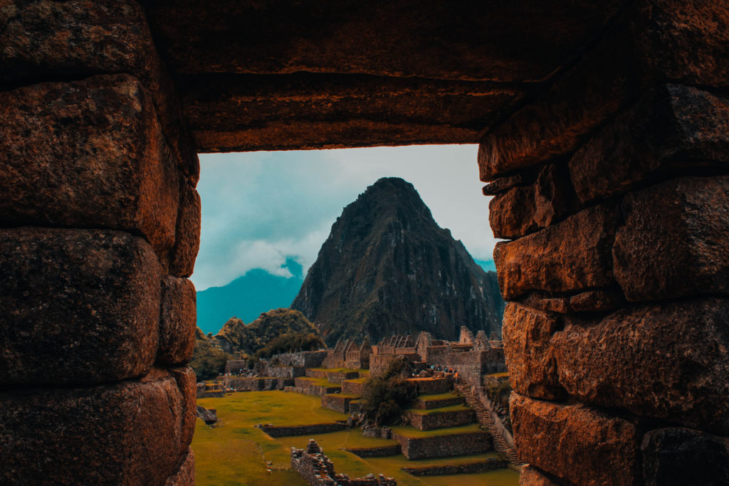 The ancient Inca citadel of Machu Picchu viewed through a stone doorway, Huayna Picchu mountain rising dramatically behind the terraced ruins under a moody, cloud-streaked sky, Peru.