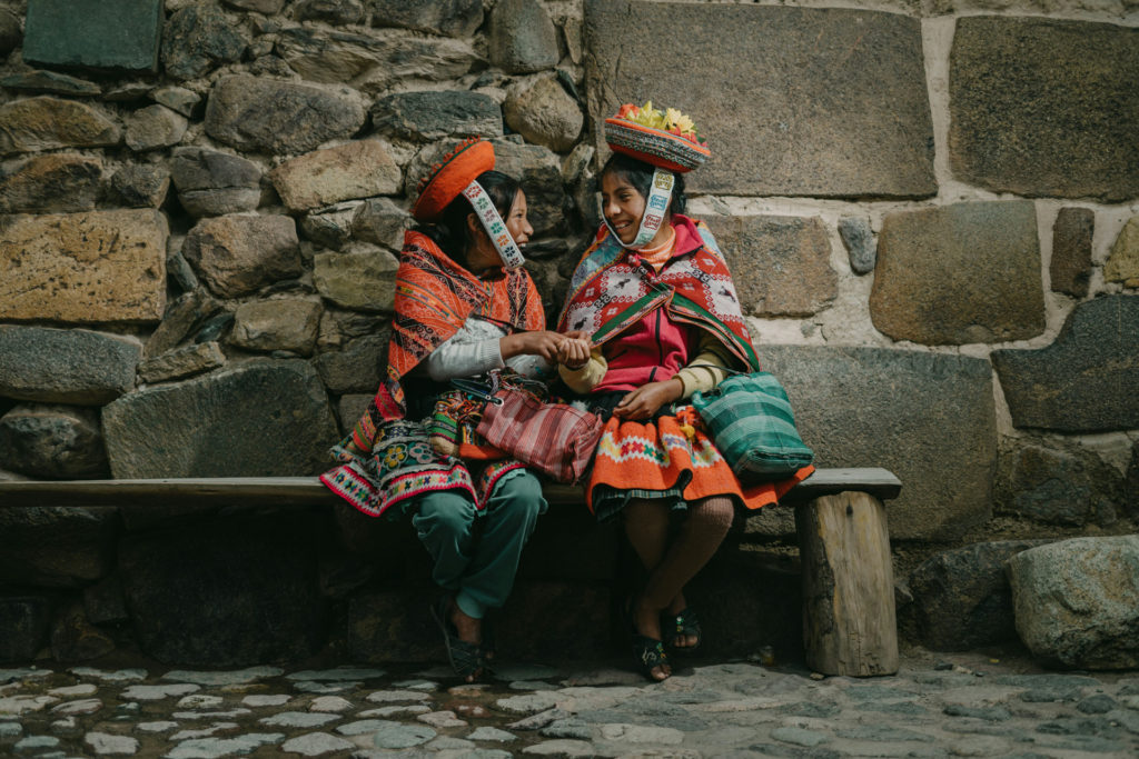 Two women in colourful traditional Andean dress and decorated hats sit together on a wooden bench against a stone wall, laughing and talking in the cobblestone streets of Ollantaytambo, Peru.