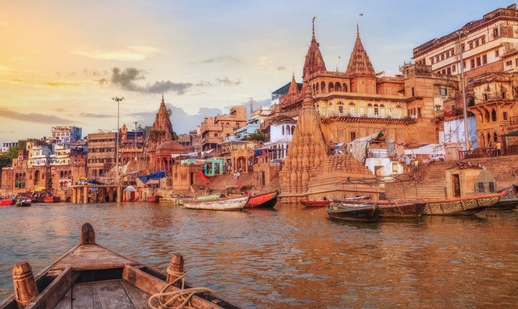 The ancient ghats and temple spires of Varanasi lining the western bank of the Ganges at sunset, wooden rowing boats moored along the steps and the city's layered sandstone architecture glowing warm gold, viewed from the prow of a river boat, India.