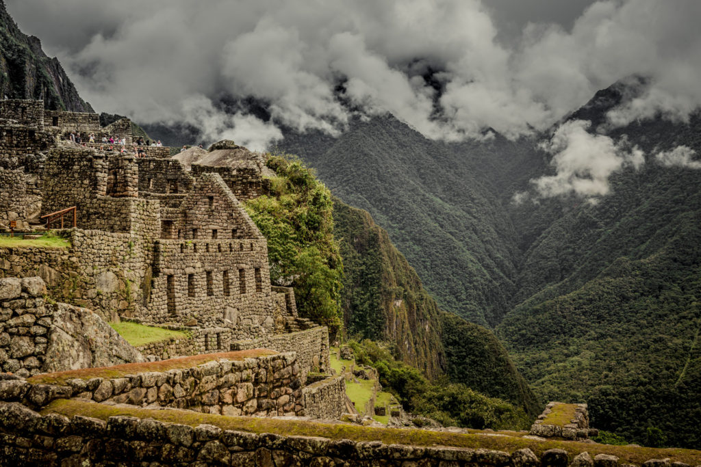 Stone buildings and trapezoidal window niches of the Machu Picchu ruins perched on the edge of a dramatic cliff, densely forested Andean mountains and rolling cloud stretching beyond, Peru.