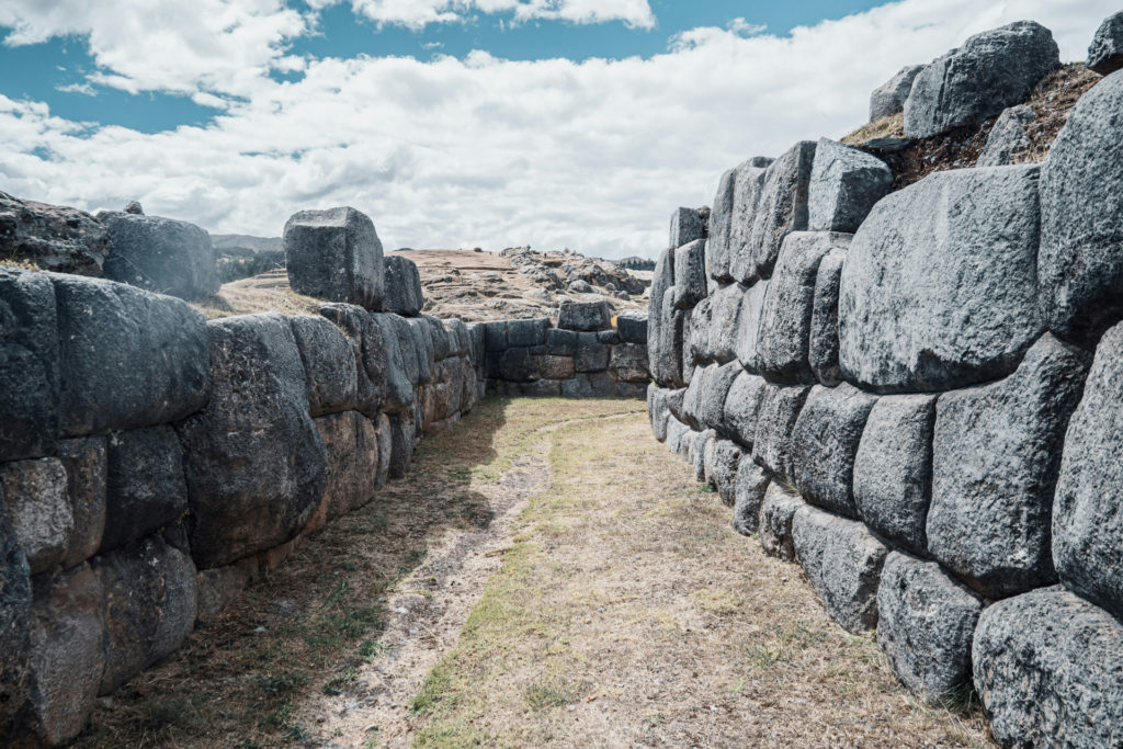 A narrow grassy passage between two towering walls of precisely fitted granite blocks at Sacsayhuamán, the enormous stones interlocking without mortar under a bright, cloud-filled sky, Cusco, Peru.