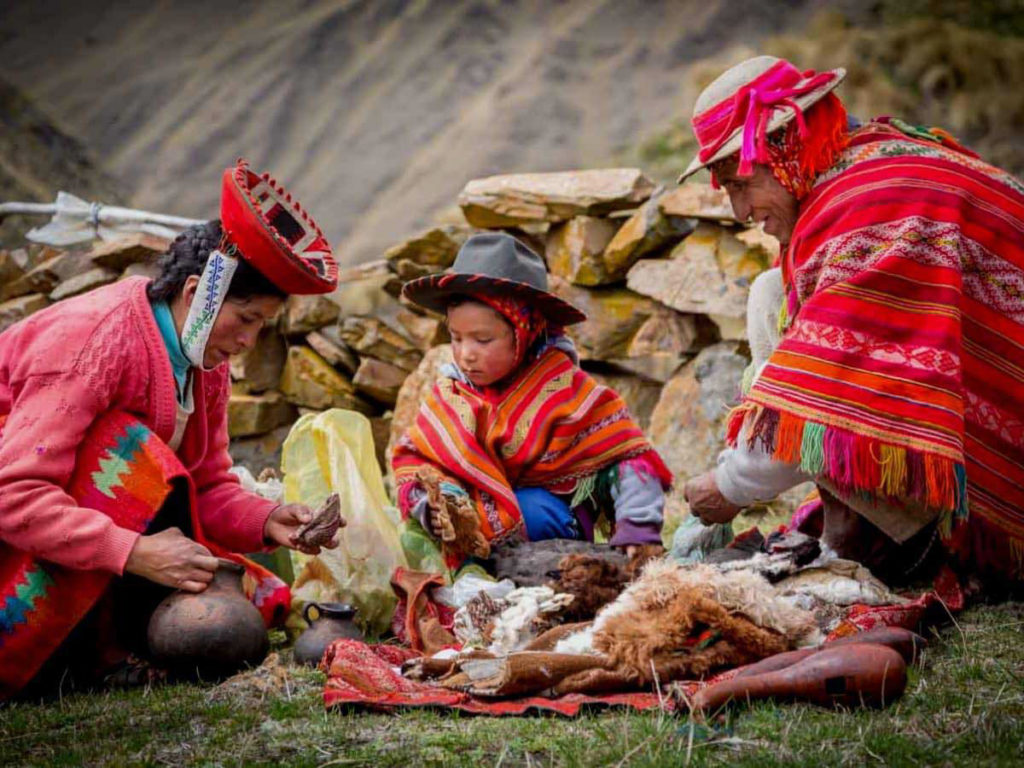 A family in vibrant traditional Andean dress sit together on an open hillside working with animal hides and natural materials, rocky stone wall behind them, Sacred Valley, Peru.