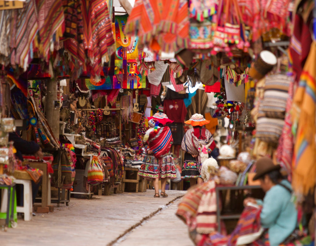 Two women in vibrant traditional Andean dress and wide-brimmed hats walk through a narrow alley bursting with colourful woven textiles, ponchos and handicrafts at the market in Pisac, Sacred Valley, Peru.