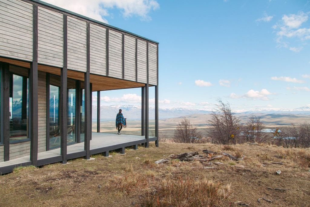 A guest wrapped in a blanket stands on the elevated timber deck of a remote lodge, gazing out across a vast open steppe, snow-capped Andean peaks and a shimmering lake stretching to the horizon, Patagonia.