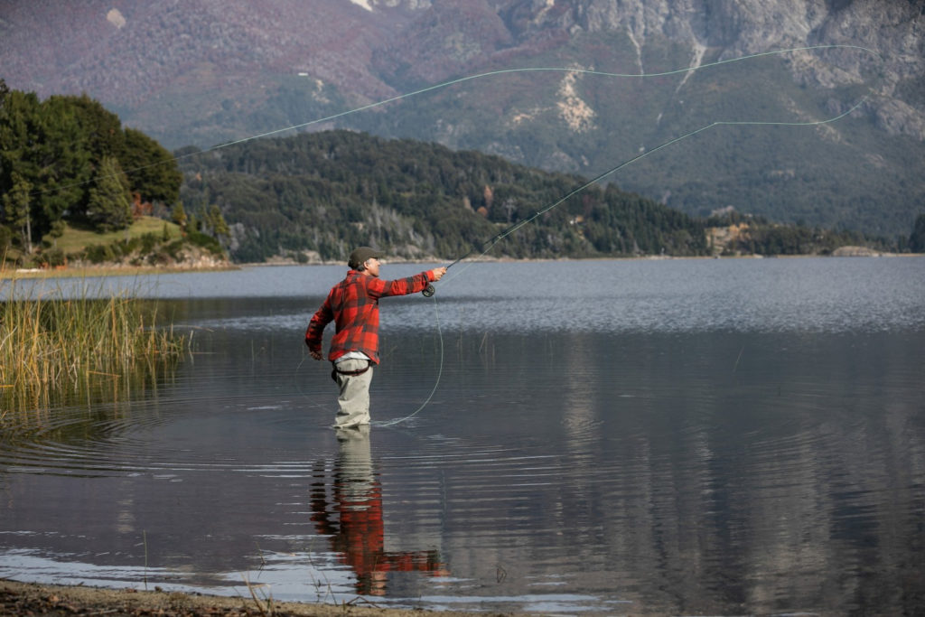 A fly fisherman in waders and a red checked shirt casts his line across a glassy Patagonian lake, forested hills and snow-dusted mountains reflecting in the still water behind him, Argentina.