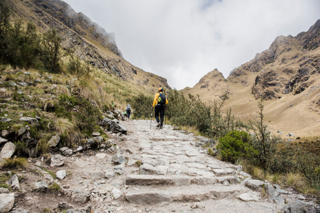 A group of trekkers ascending ancient stone steps on the Inca Trail, a dramatic high-altitude mountain pass with bare peaks and a pale sky opening ahead, Peru.