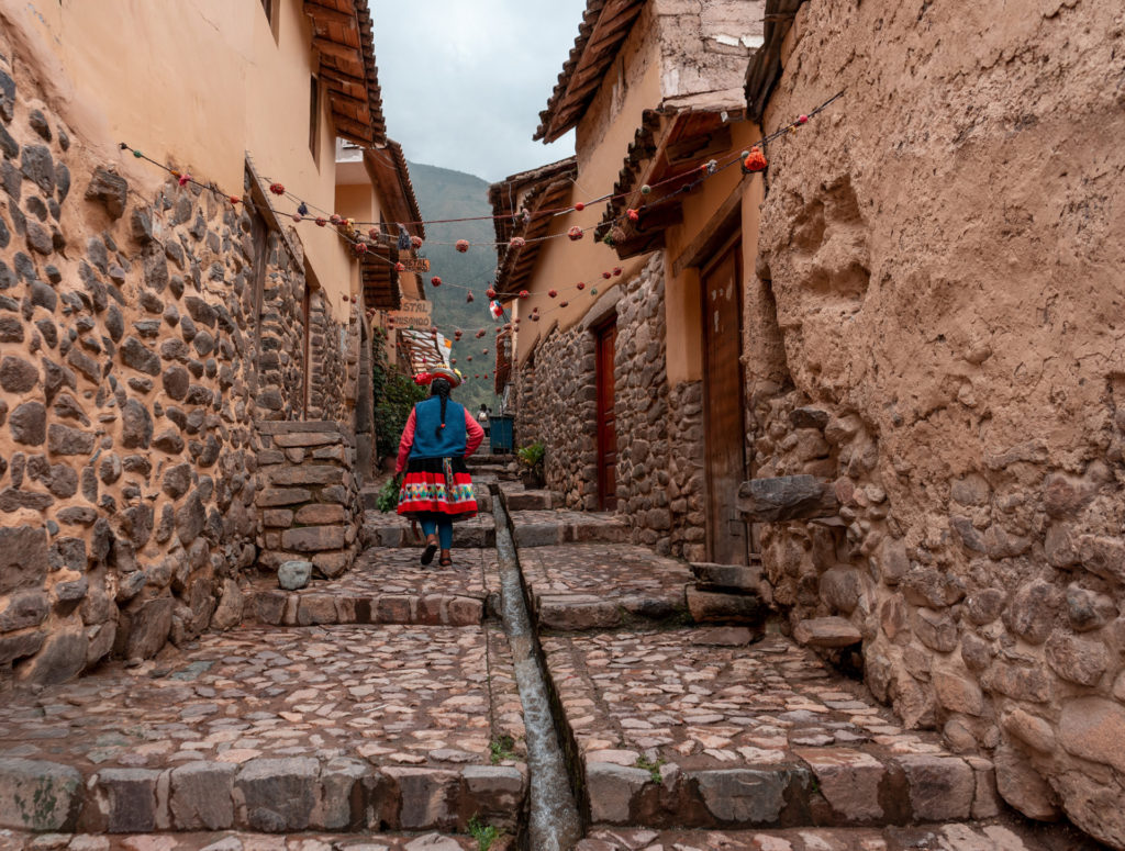 A woman in traditional Andean dress and a flower-adorned hat walks up a narrow cobblestone lane with a central water channel, colourful bunting strung between stone and adobe walls, Ollantaytambo, Peru.