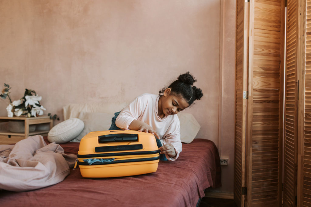 A young girl kneels on a bed, eagerly zipping open a bright yellow suitcase, a warm-toned bedroom with timber louvred doors and a bedside plant visible behind her.
