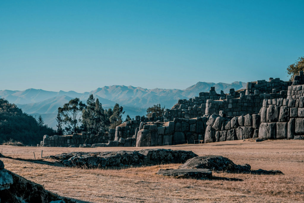 The massive zigzagging stone walls of the Inca fortress of Sacsayhuamán above Cusco, enormous fitted granite blocks stretching across a dry grassy plain with the Andean mountain range under a clear blue sky beyond.