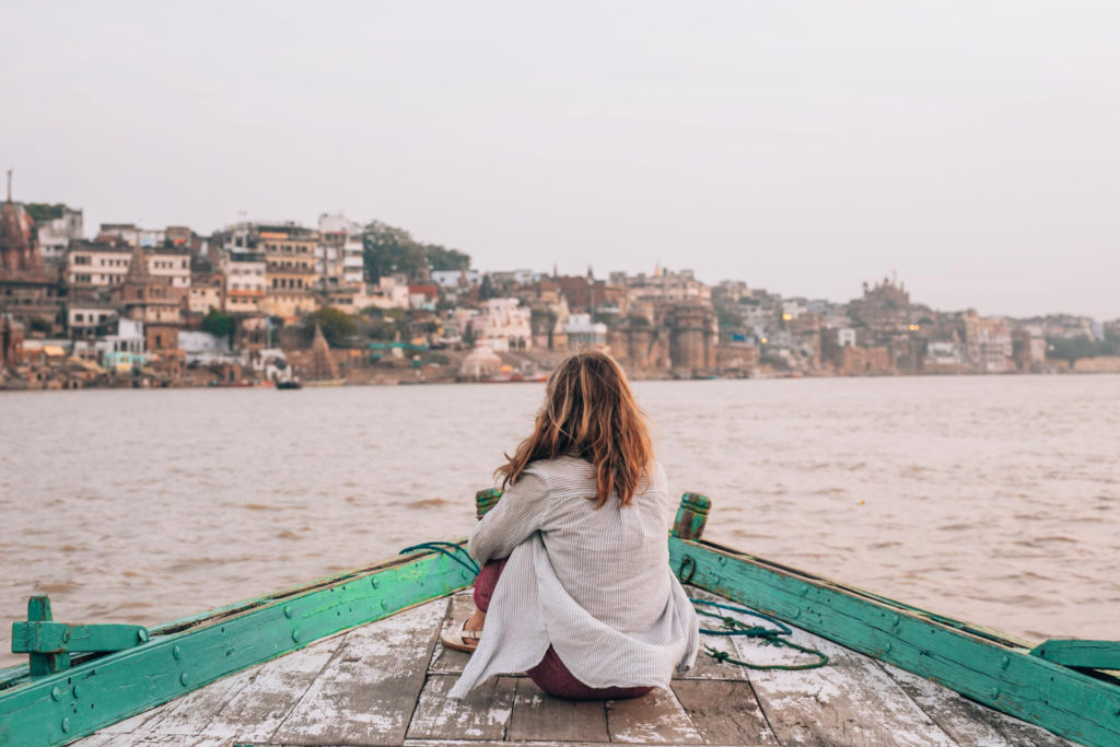 A woman sits alone on the bow of a wooden river boat, gazing toward the ancient ghats and temples of Varanasi rising from the far bank of the Ganges in soft morning haze, India.