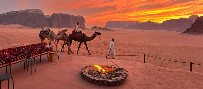 Camels and Bedouin guides move quietly through the desert as the sun sets behind Wadi Rum’s monumental cliffs.