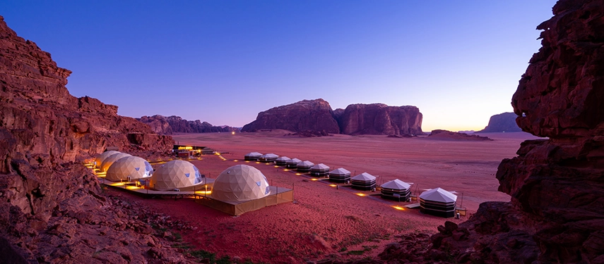 At dusk, softly lit desert domes and tents glow beneath a deepening sky, surrounded by the vast stillness of Wadi Rum.