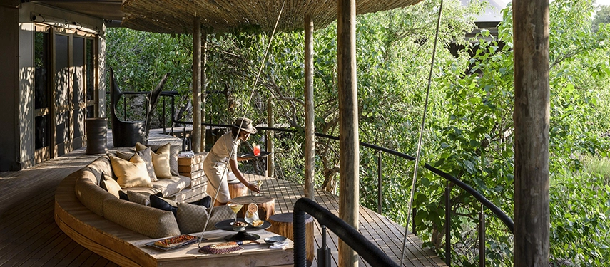 A staff member serves drinks on the shaded timber deck of a Xigera suite, surrounded by the dense green canopy of the Okavango Delta, with deep curved sofas and scatter cushions in the foreground.