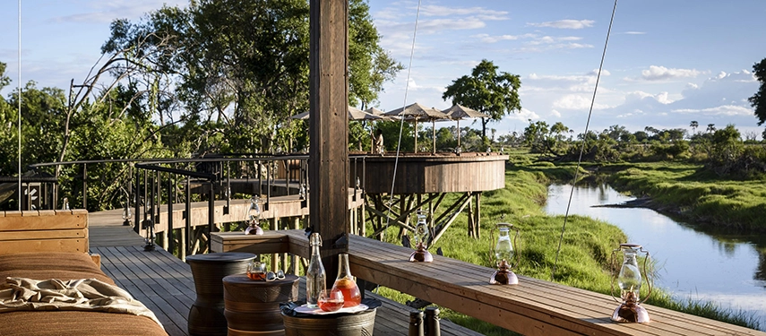 The timber viewing deck of Xigera Safari Lodge looking out over a green Delta channel, with drinks set on barrel tables, copper lanterns catching the light, and a raised tented deck visible across the water.
