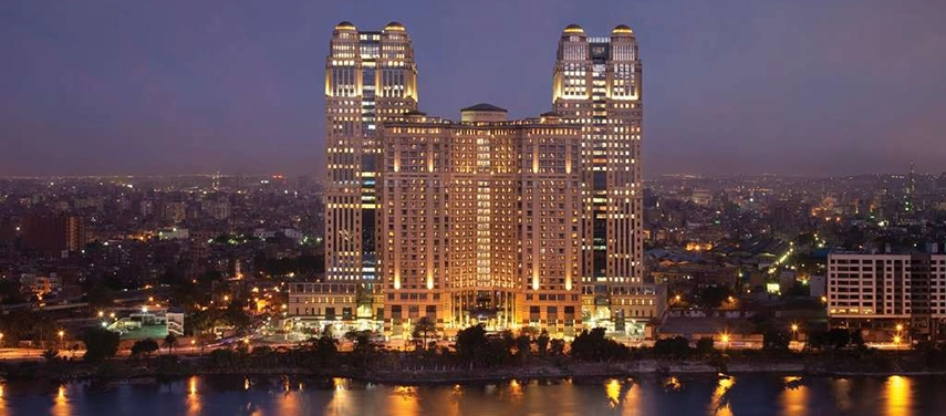 The Sofitel Cairo Nile Hotel towers glow against the evening sky, reflecting across the Nile with Cairo’s city lights stretching into the distance.