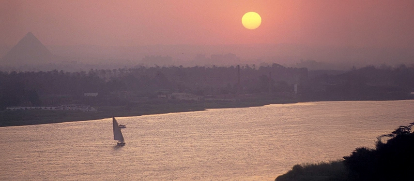 A lone felucca sails along the Nile at sunset beneath a hazy sky with the silhouette of a distant pyramid.