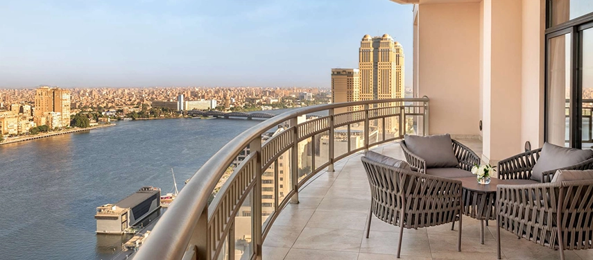 A spacious balcony with woven chairs overlooks the Nile and Cairo’s skyline under clear afternoon light.