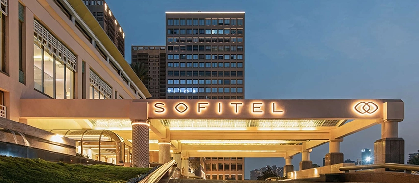The Sofitel Cairo Nile Hotel’s illuminated entrance glows warmly against the evening sky with the city skyline rising behind it.