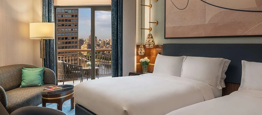 A contemporary guest room at Sofitel Cairo Nile Hotel offers plush bedding and a private balcony overlooking the river and city skyline.