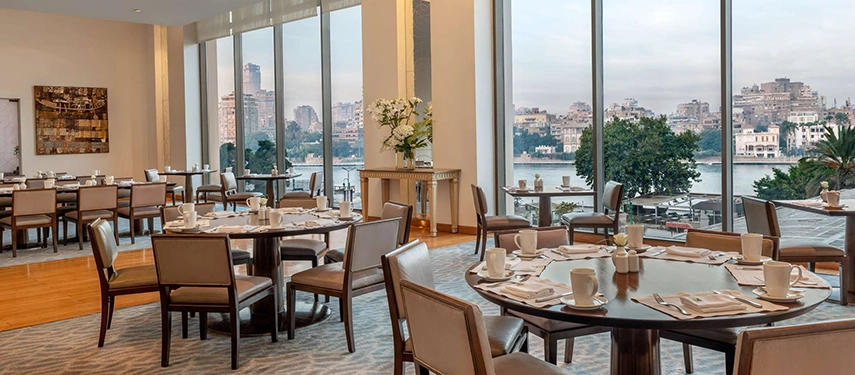 A bright dining room with floor-to-ceiling windows looks out toward the Nile, creating a serene riverside setting for meals.