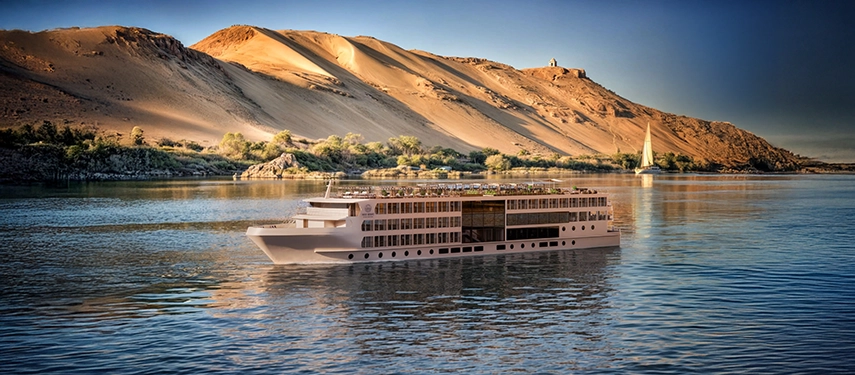 A&K Sanctuary Nile Seray cruising the Nile beneath golden desert dunes, with calm waters reflecting the light and a traditional felucca sailing nearby.