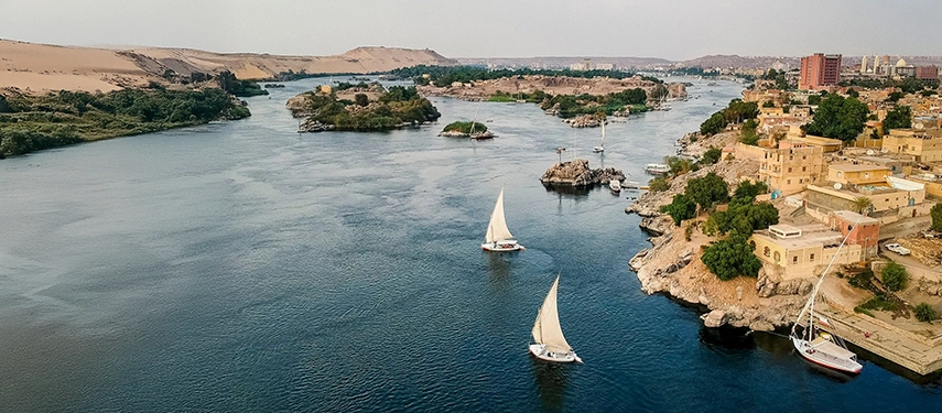 Aerial view of the Nile winding past desert hills and riverside towns, with traditional feluccas sailing through calm blue waters.