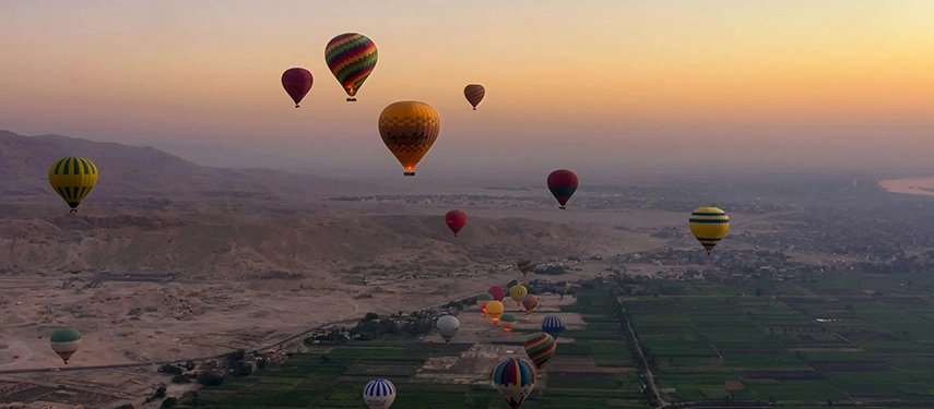 Hot air balloons floating above the Nile Valley at sunrise, drifting over green farmland and desert plains near Luxor.