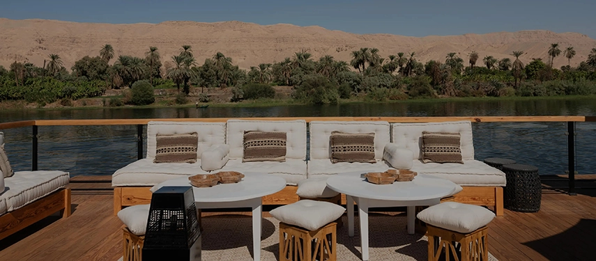 An open-air lounge deck aboard A&K Sanctuary Nile Seray, furnished with cushioned seating and river views framed by palm-lined banks.