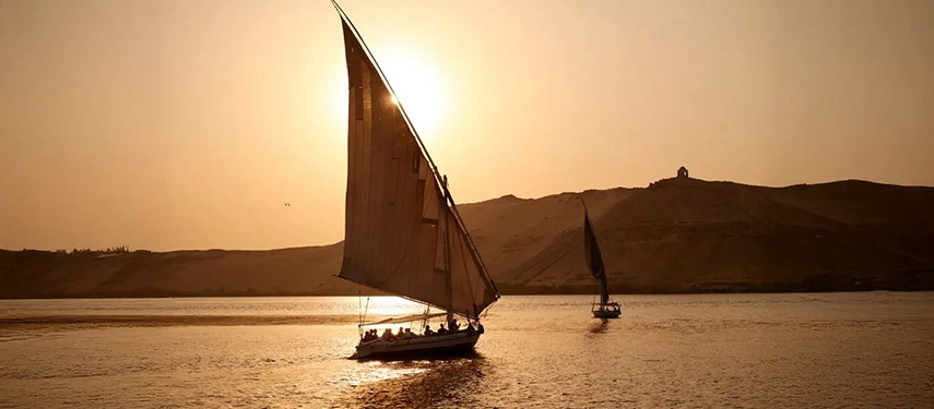 Traditional feluccas sailing across the Nile at sunset, silhouetted against golden light and desert hills beyond.