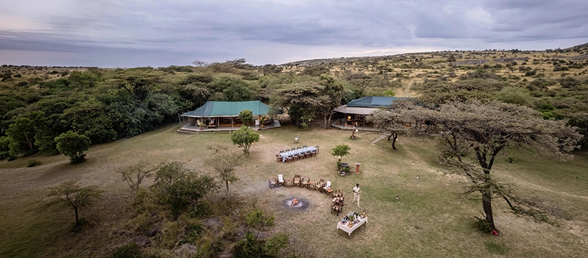 An aerial view of Hemingways River Camp Mara showing the main camp buildings, an outdoor dining table set for dinner and a fire circle arranged on the open lawn, surrounded by the Mara bush.