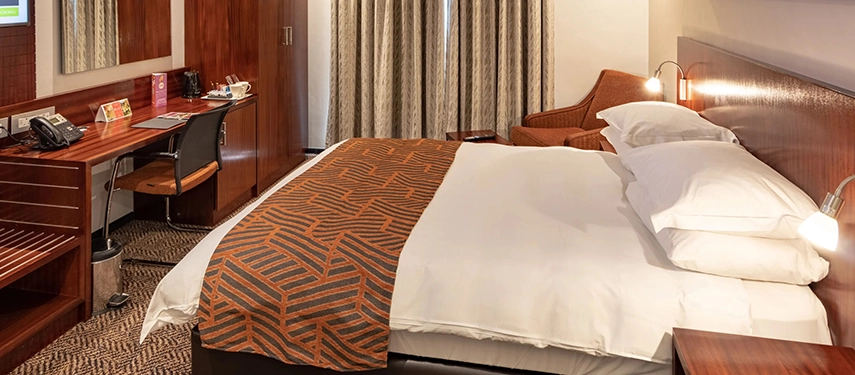 A welcoming and functional guest room offering a plush bed, rich timber furnishings, and a dedicated workspace for catching up before a flight.